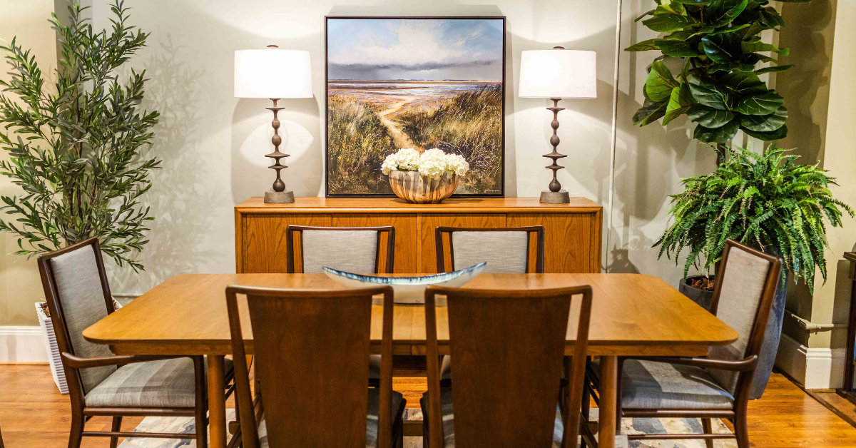 A wooden dining table with 6 chairs around it and a wooden stand with two lamps, a piece of artwork, and a bowl with flowers on top all in between two plants