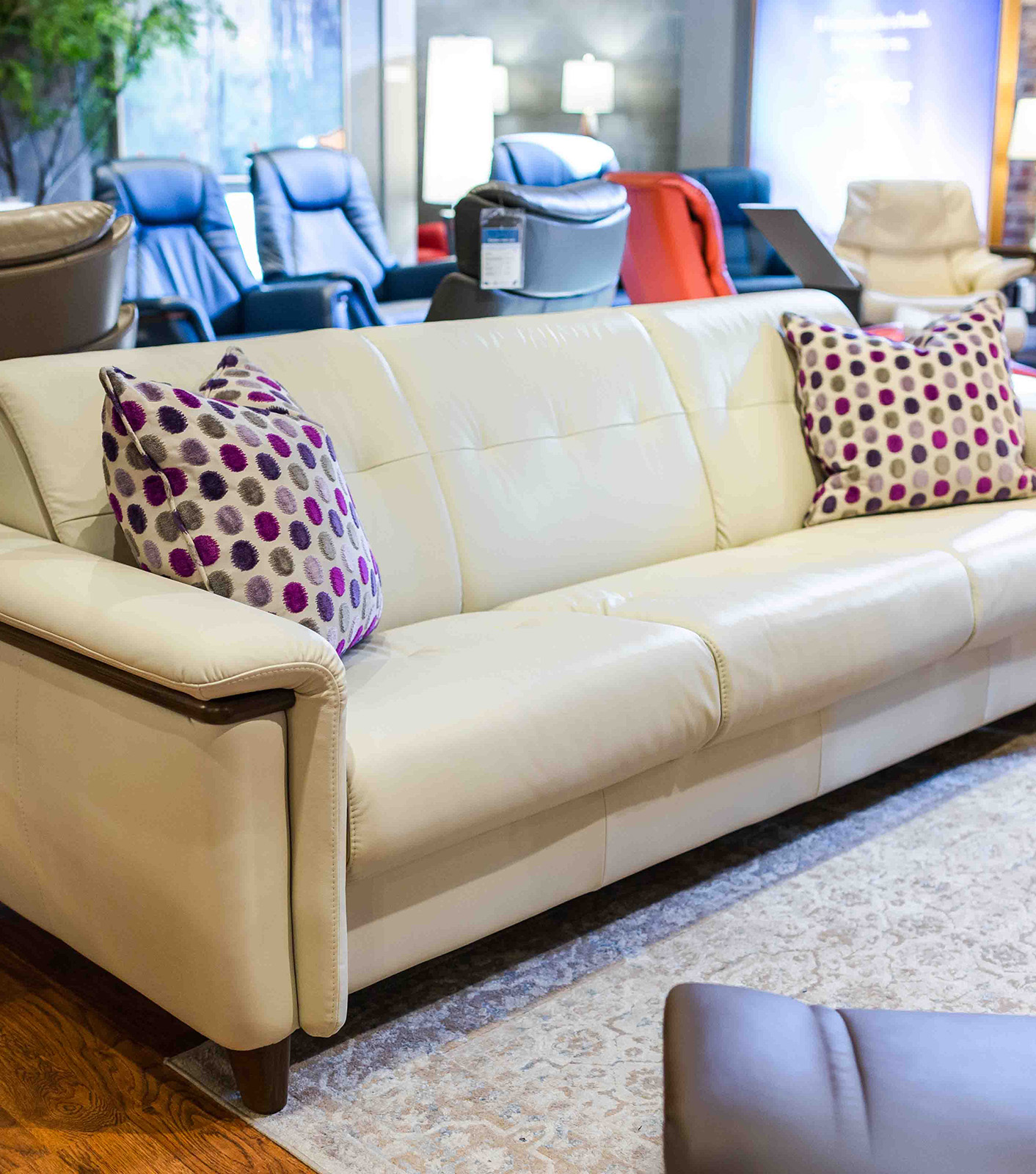 A light colored couch with a pillow with purple and grey polka dots in our Roanoke Furniture Store. Two leather like material chairs in the background.